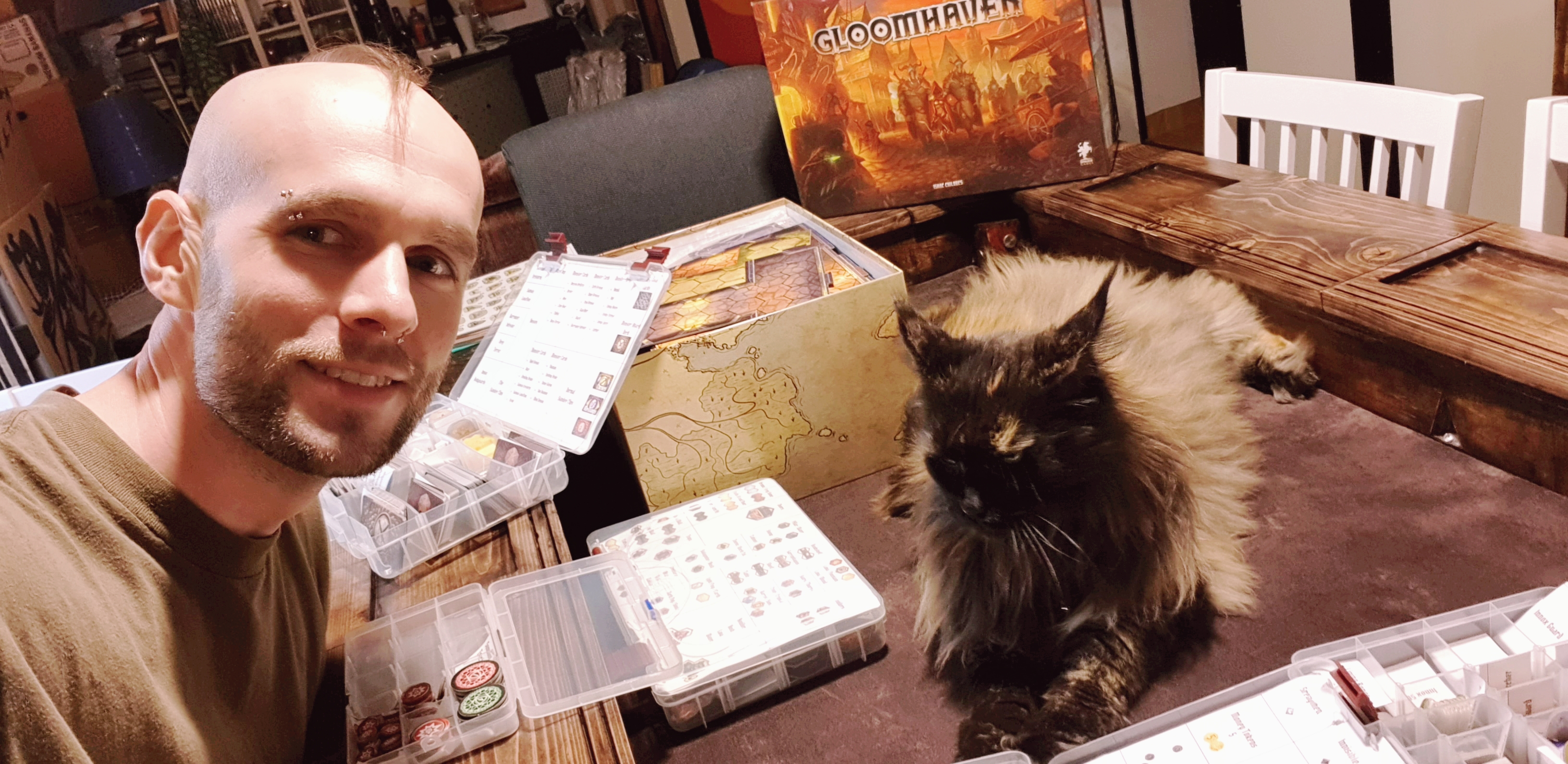 Christian taking a photo of himself smiling and playing the board game Gloomhaven with his cat Lola laying across the table.